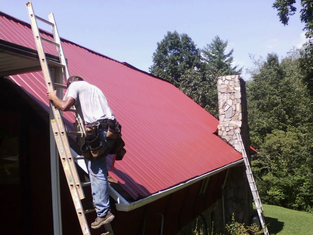 Roofer on a ladder beside a red metal roof and stone chimney — Soffit & Fascia Repair by Roofing Brothers in Castle Creek
